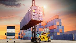 A forklift lifting a shipping container at a port during sunset, with stacked containers and a truck in the foreground.